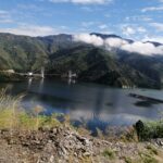 A landscape with the view of a lake, across which is a mountain range. And one of those mountain is covered with bright white clouds.